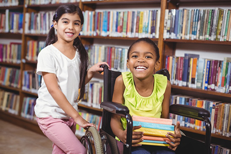 4.1.1 LearnExplore_Books_RC_Kids Child stands with a friend who is in a wheelchair while they both enjoy looking for books at the library (Links to kids booklist)