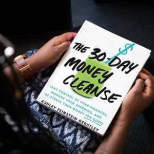 A woman sits with the book "The 30-Day Money Cleanse" on her lap. The cover includes a white background with a green smoothie and a dollar sign straw.