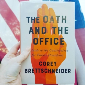 A book with an orange hand under the title The Oath and the Office is held up in front of an American flag.